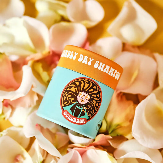 A turquoise blue and orange topped cardboard tube of Scrubber Ylang & Rose Dry Shampoo. The tube is sitting on a blurry collection of pink and cream flower petals.