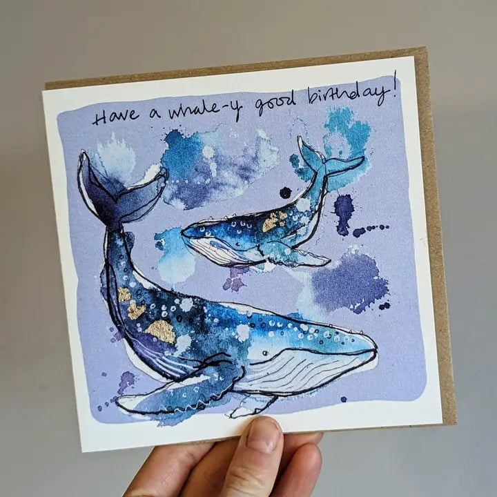 Tori Ratcliffe Art Hump Back Whale card with black writing that says “Have a Whale-y Good Birthday”. The watercolour whales contain blues, purples, white and gold, and the background of the card is very light purple. There is a bigger whale in the foreground and a slightly smaller whale behind. The card is being held by a hand at the bottom, has a brown envelope sticking out of the right hand side and it is on a white background.