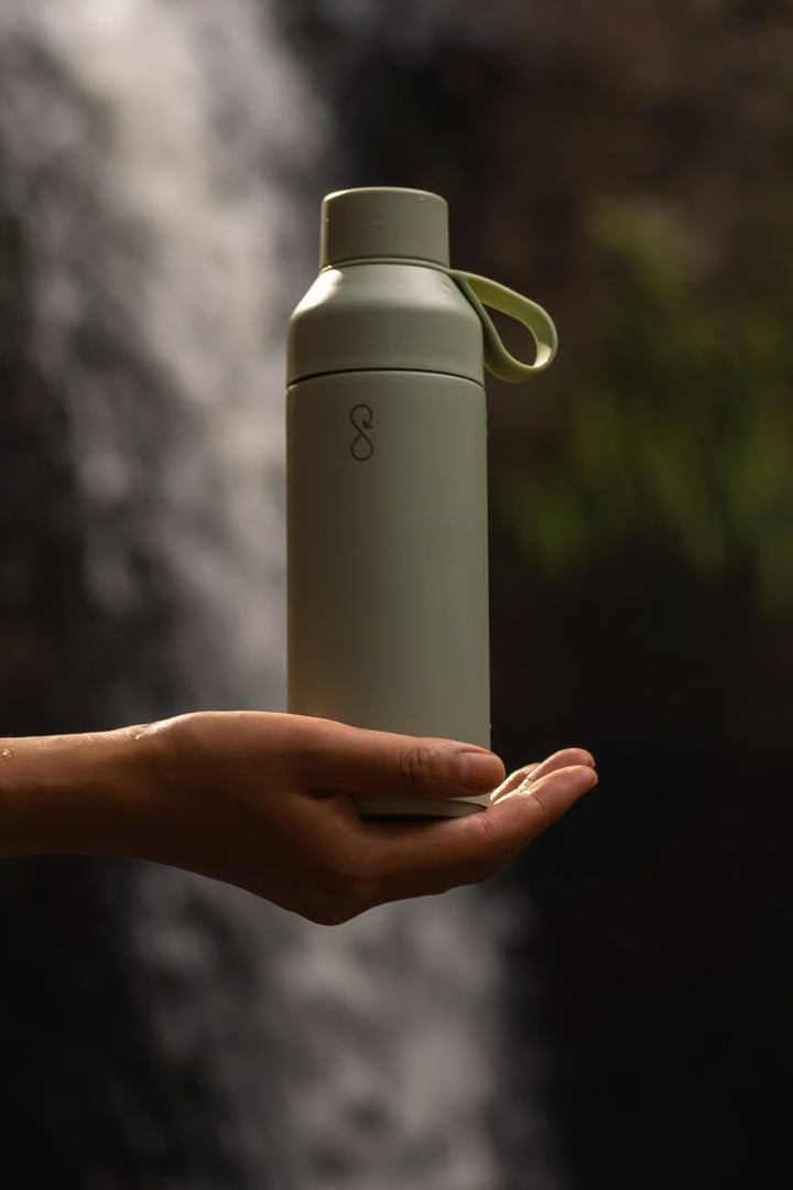 Ocean Bottle Shale Green 500ml Bottle balances on a hand coming from the left. There is a waterfall in the background.