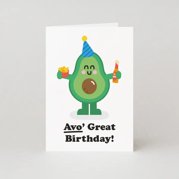 Studio Boketto birthday card with a cartoon like celebrating avocado. The green avocado has a blue party hat on, and is holding a beer in the hand to the right and a snack in the hand to the left. ‘Avo great birthday’ is written in black underneath the avocado, with ‘Avo’ being underlined. The card is white and is stood in a white background.