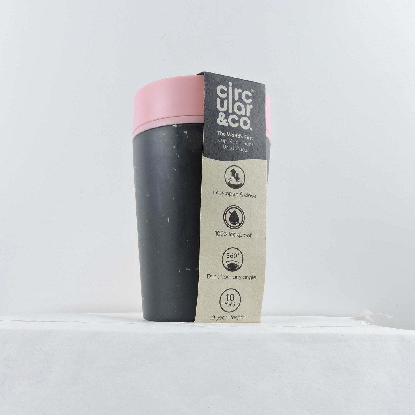 Circular & Co 8oz Black cup with a pink lid. It has a card beige coloured sleeve (with a dark grey top edge) over the top of it detailing information about the cup. It is sat on a white background.