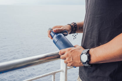 Ocean Bottle Ocean Blue 500ml Water Bottle being held by a man in a black top with a watch on one arm and bracelets on the other. He is stood to the right and is holding the bottle (centre) near a metal railing, with the ocean in the background.