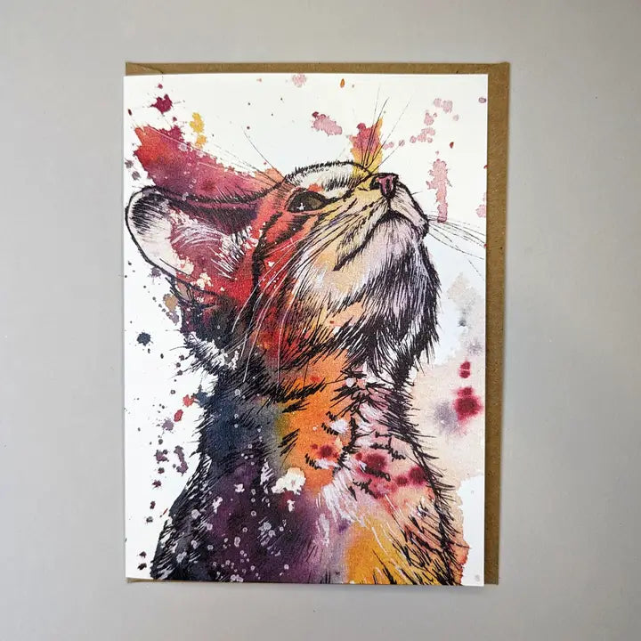 Tori Ratcliffe Art watercolour and pen drawing of a kitten. The kitten is looking up to the right top corner of the card and is coloured with patches and blends of purples, pinks, very dark blues and yellows. The card has the edge of a brown envelope sticking out the right side and it is placed on a white background.