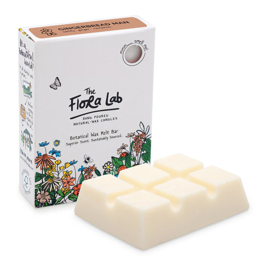 The Flora Lab Gingerbread Man Scented Wax Melts. A slab of white wax melts, unboxed in the foreground, with the white box (with floral decoration around the bottom) stood on end slightly behind and to the left.
